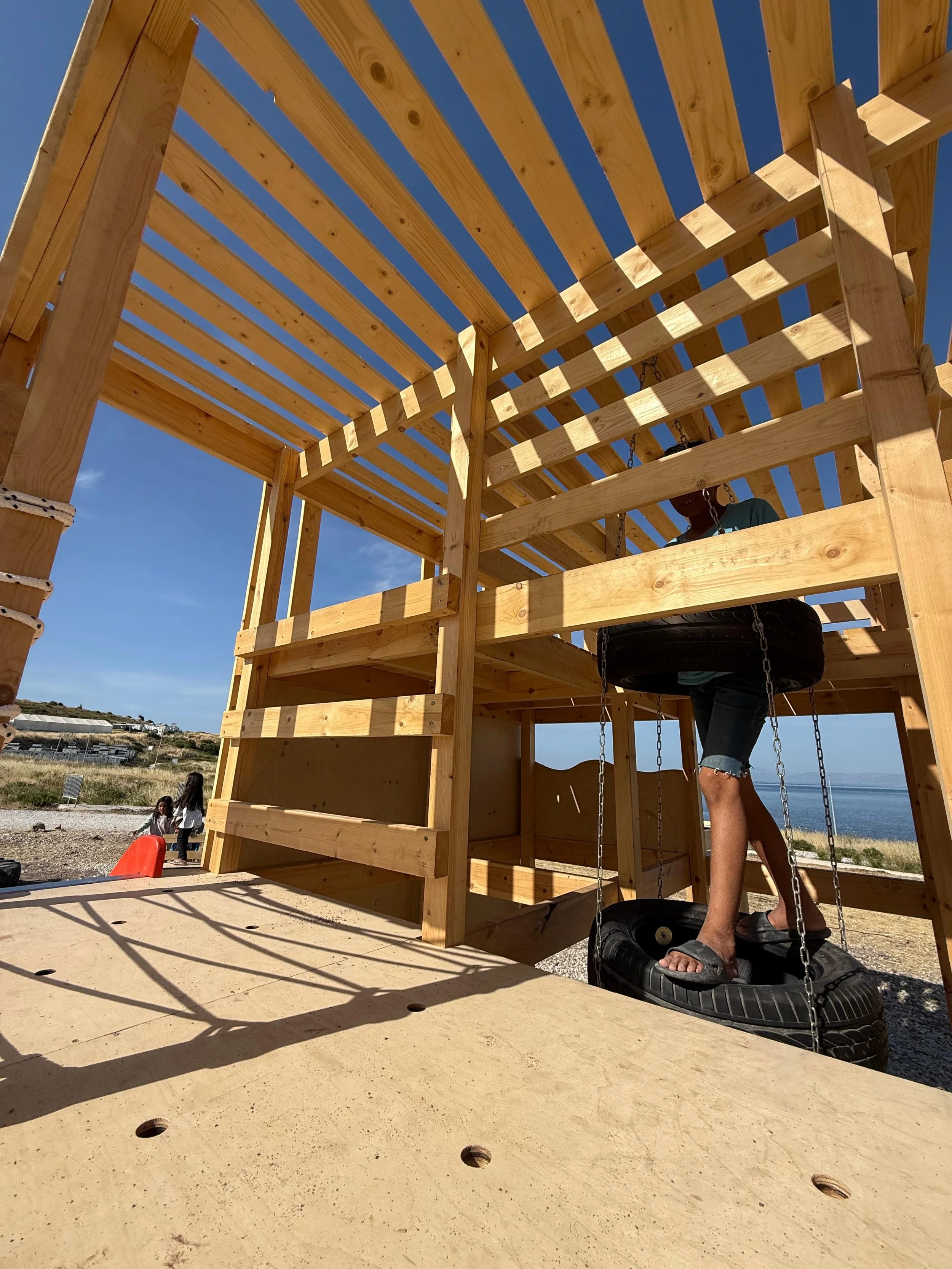 A wooden play structure with a tire swing hanging inside, located outdoors near a beach. There are children and adults in the background, under a blue sky.