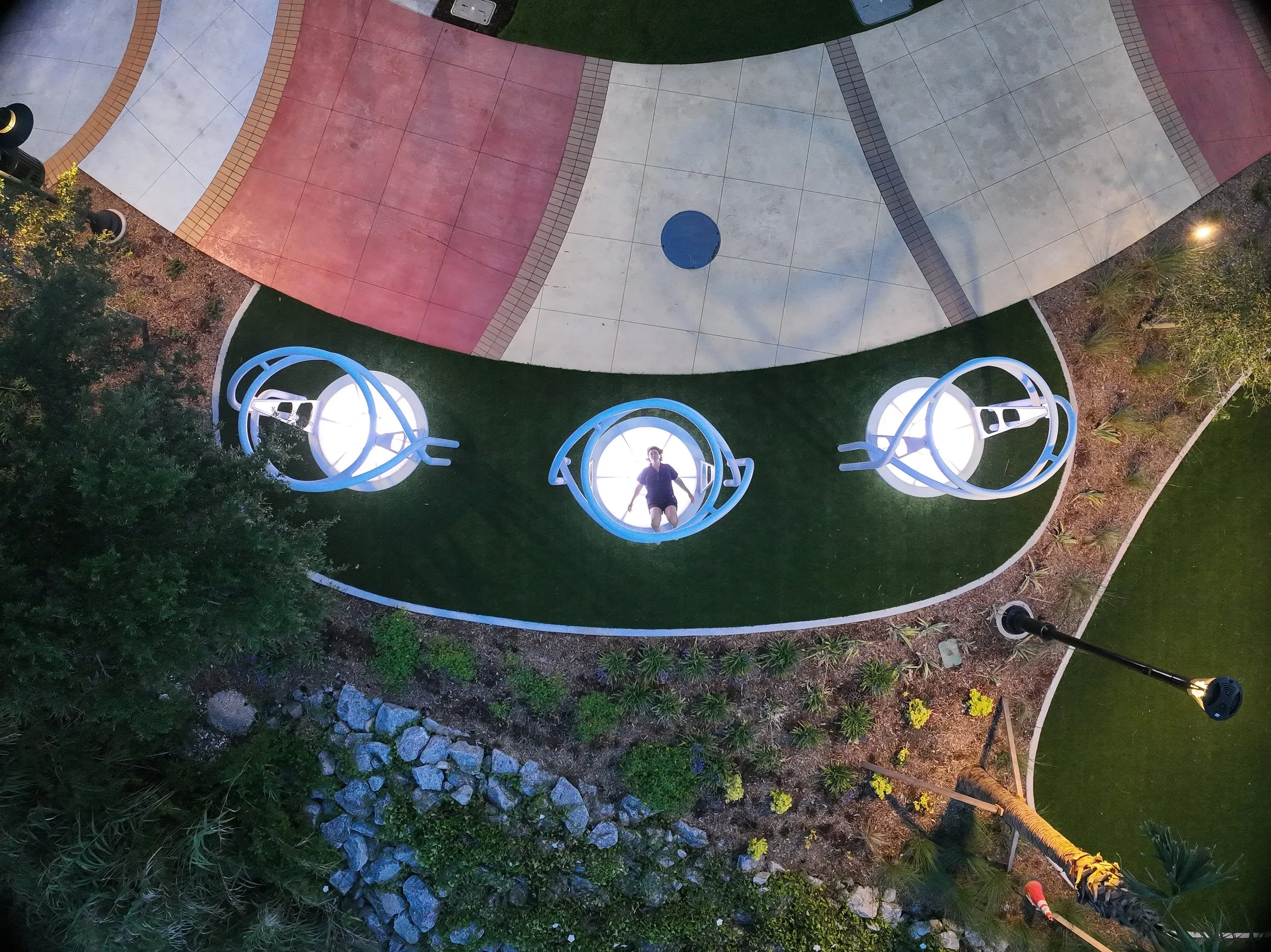 An aerial view of a colorful park with curved paved walkways in red, gray, and white, a blue circular sculpture with a person sitting in it, green grass, trees, and outdoor lighting at dusk.