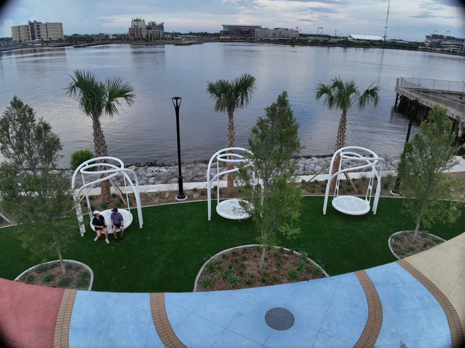 A riverside park with three white swing chairs, two people sitting on one of them, surrounded by green trees, palm trees, and a path with colorful paving stones. In the background, there is a calm river and city buildings.