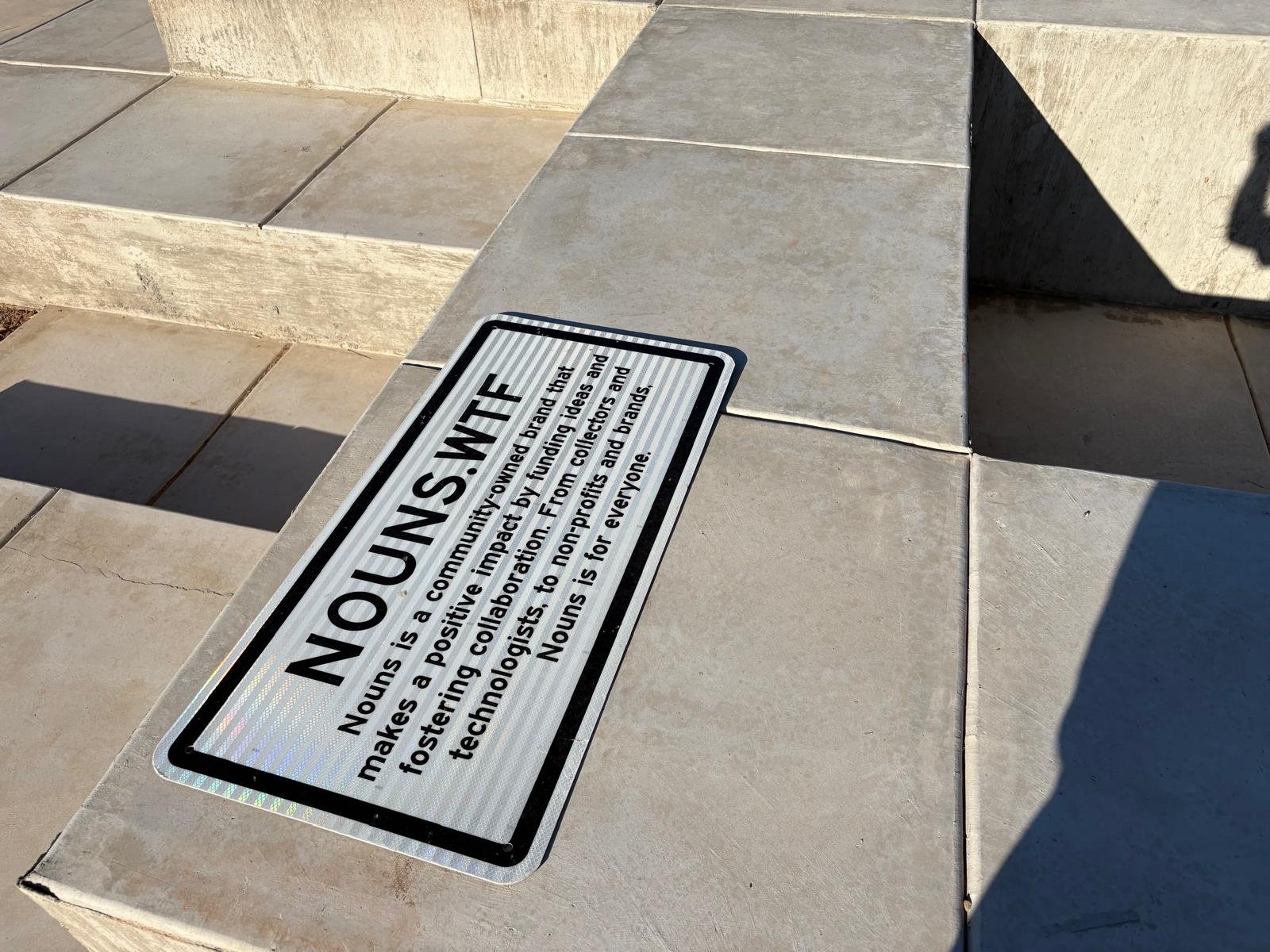 Text-based sign on a concrete surface explaining the mission of a nonprofit called NOUNS. N.T. It highlights positive impact by funding ideas and fostering collaboration to benefit everyone.