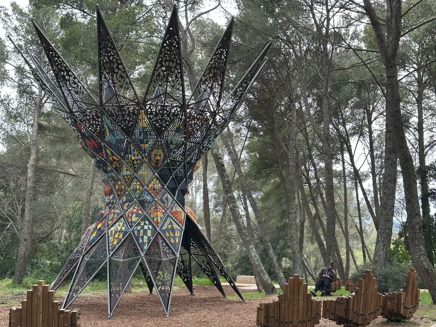 A colorful, modern, geometric sculpture in a wooded park, with trees surrounding it and a person sitting on a bench nearby.