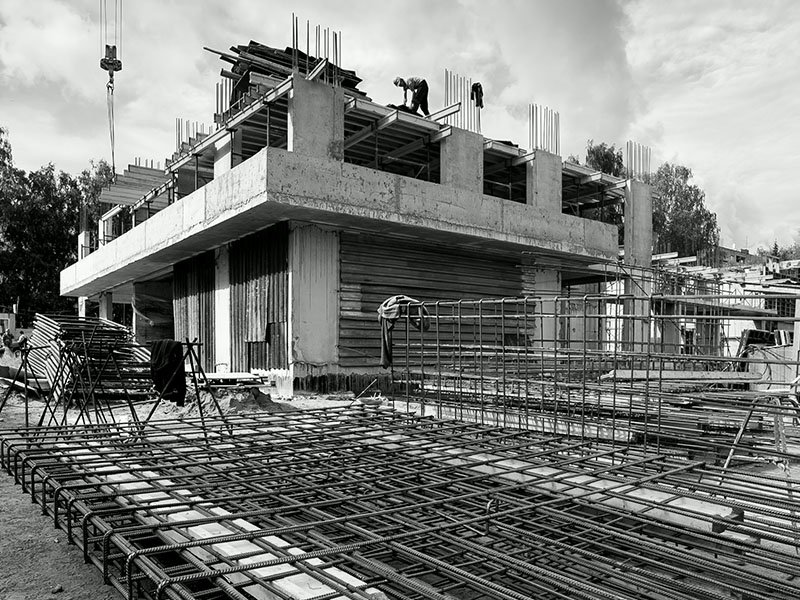Construction site with workers on upper floor of building under construction, concrete and steel framework visible, steel reinforcement bars in foreground.