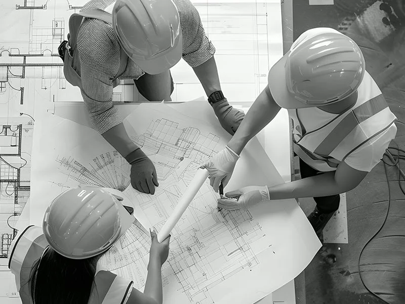 Three construction workers wearing helmets and gloves working on blueprints at a table.