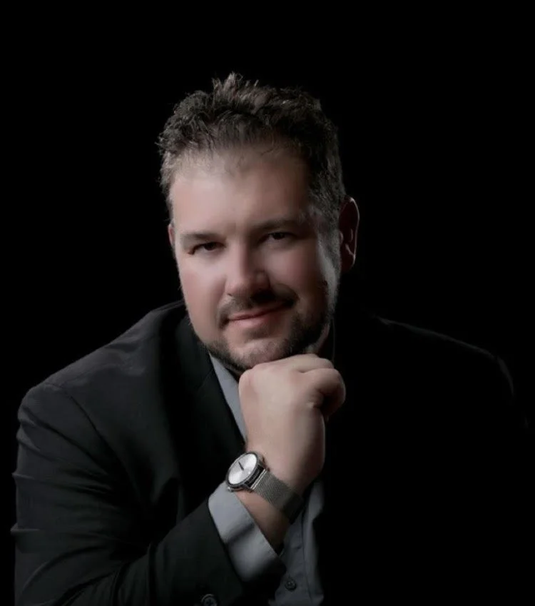 Portrait of a man with tousled hair in a dark suit, resting his chin on his hand, against a black background.