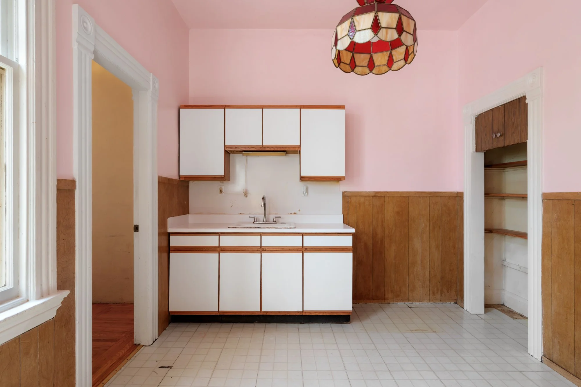 Room with a pink wall, a small kitchen sink, white upper cabinets, and a wooden wall panel. A stained glass pendant lamp hangs from the ceiling, and there are two doorways on either side.