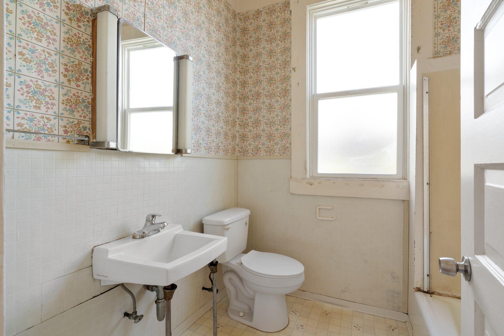 A small bathroom with a white toilet, a white sink, a mirror, and a window. The walls are decorated with patterned floral tiles and plain white tiles. The bathroom appears old and slightly worn.