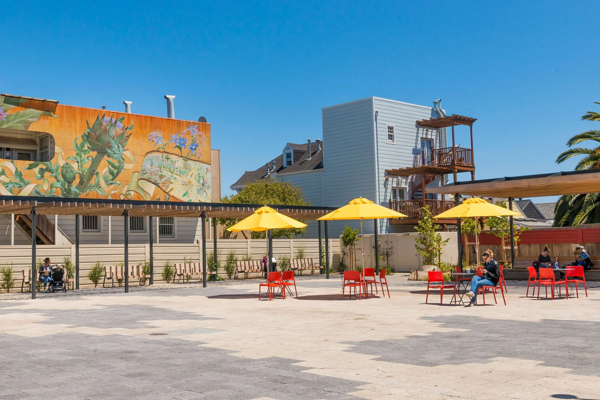 Public outdoor seating area with red chairs, yellow umbrellas, benches, and people sitting, with a colorful mural on a building and residential houses in the background under a clear blue sky.