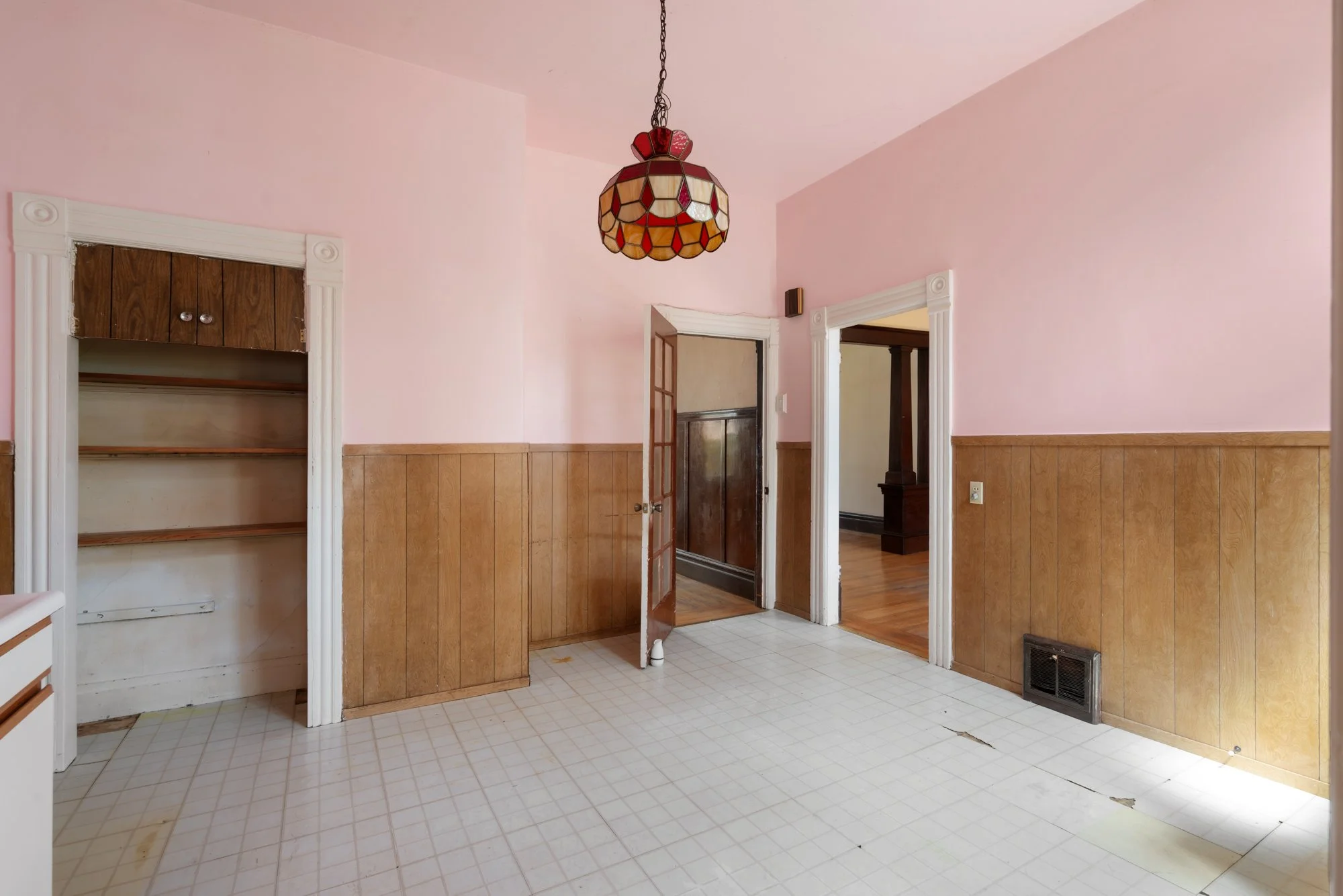 Room with pink walls and wood paneling, featuring a stained glass ceiling light, an open doorway to a dark wood floored room, an empty built-in shelf, a broken tile floor with visible damage, and a floor vent.