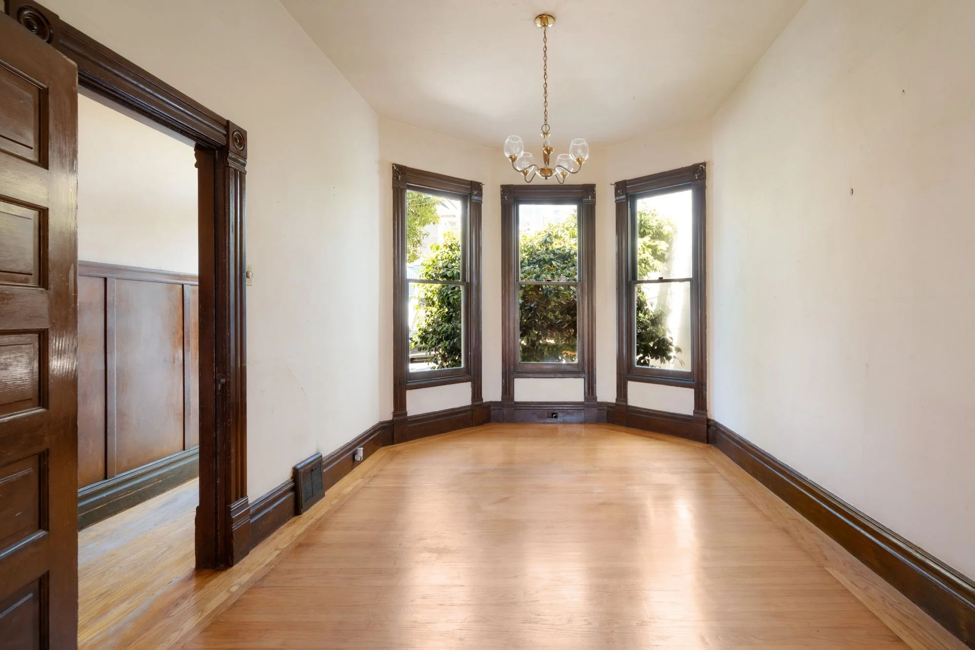 An empty room with wooden floors and three tall windows with dark wood trim, letting in natural light and showing greenery outside. A chandelier hangs from the ceiling, and there is decorative dark wood trim around the windows and doorways.