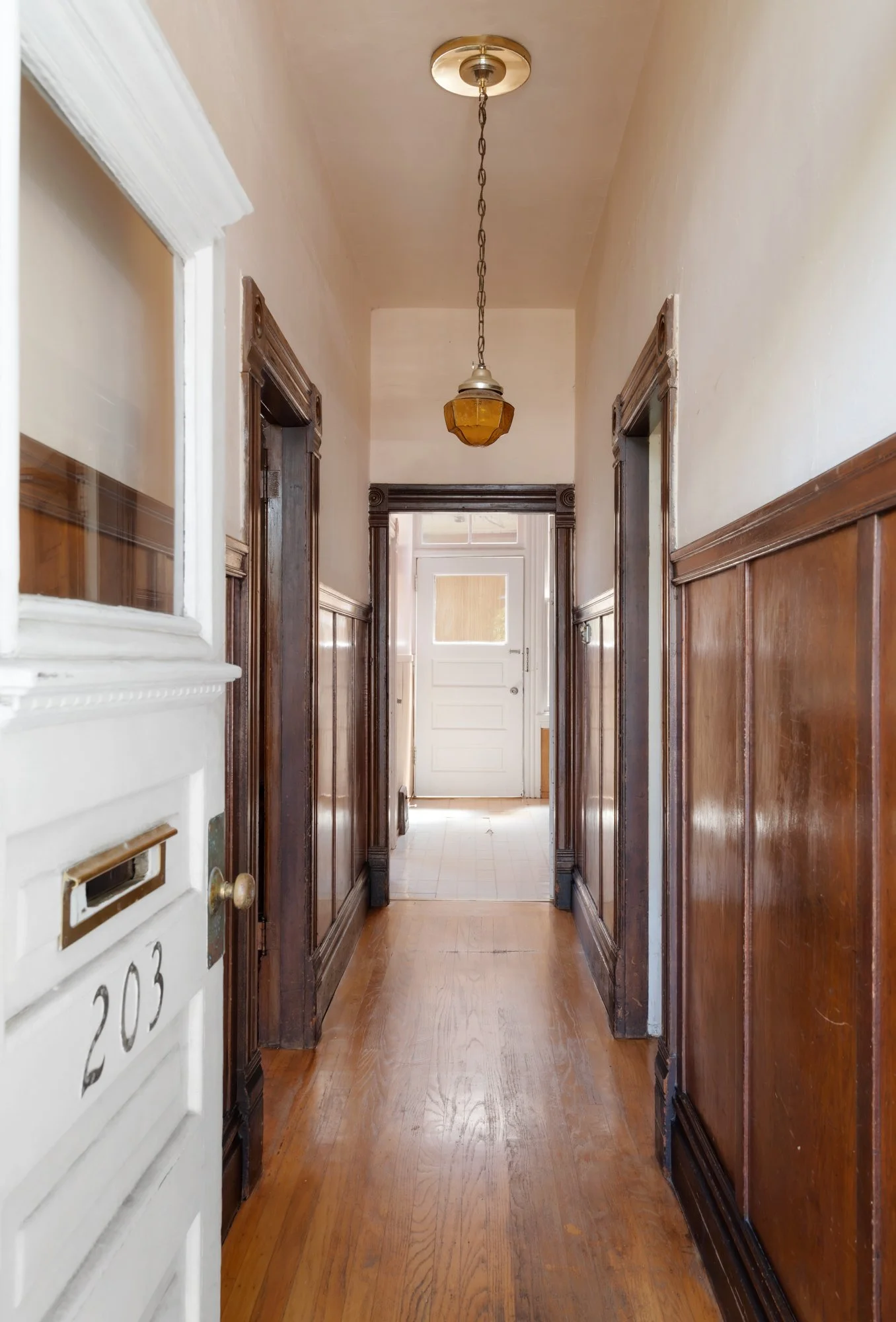View of a narrow hallway with hardwood floors, wooden wall paneling, and a vintage hanging ceiling light. At the end of the hall, a white door with a frosted window.