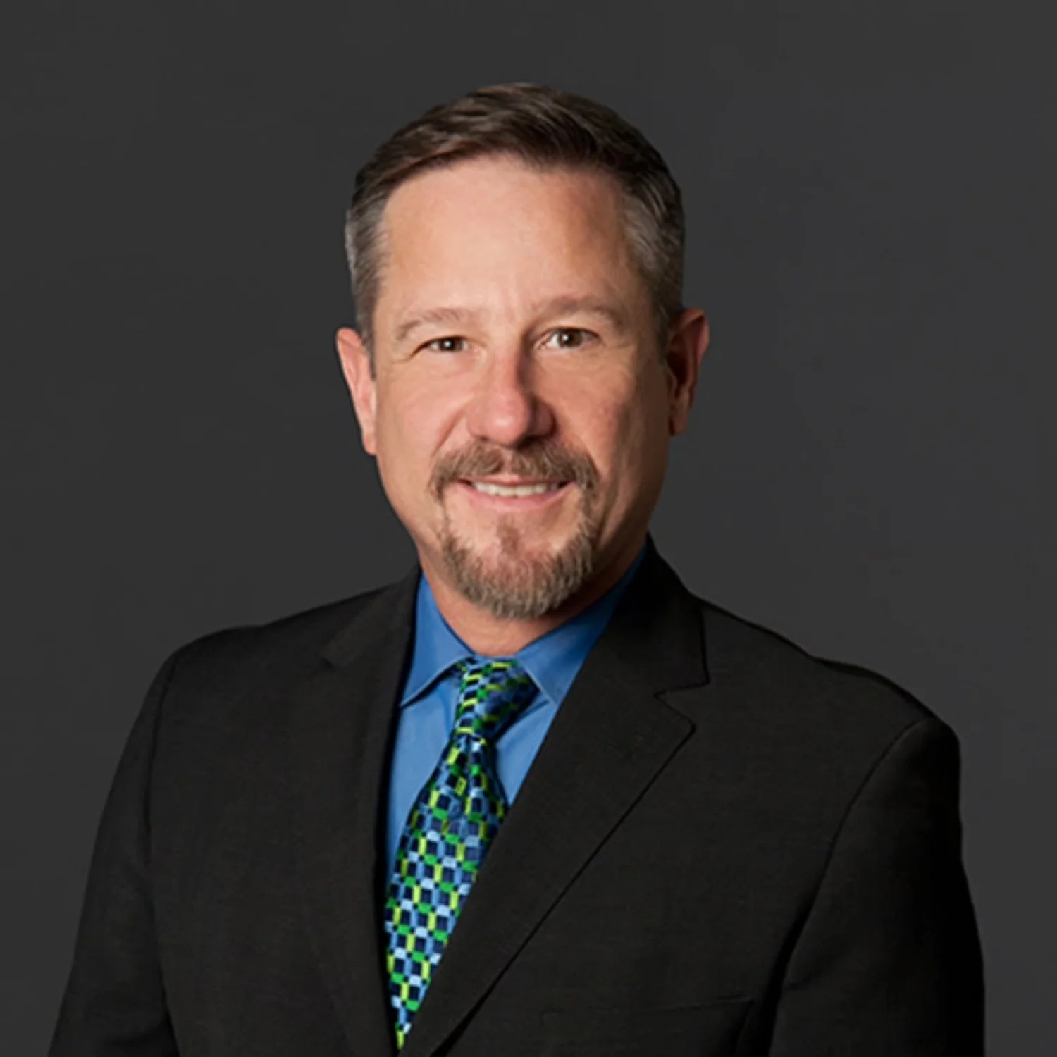 A professional headshot of a middle-aged man with brown hair and a beard, wearing a dark suit, blue shirt, and a multicolored tie, against a dark gray background.