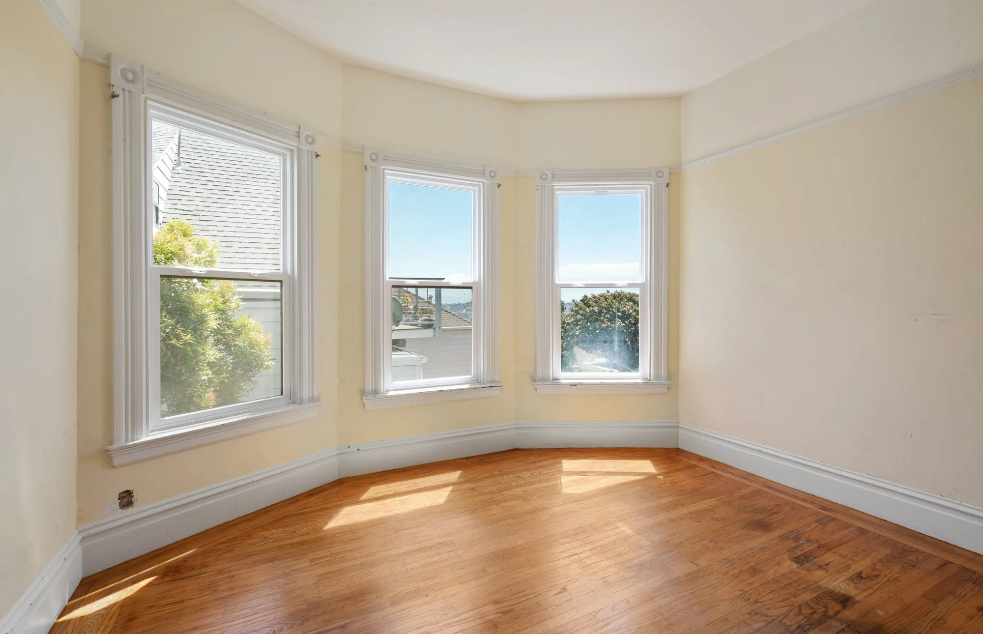 Empty room with three large bay windows, beige walls, wood floor, and white trim.