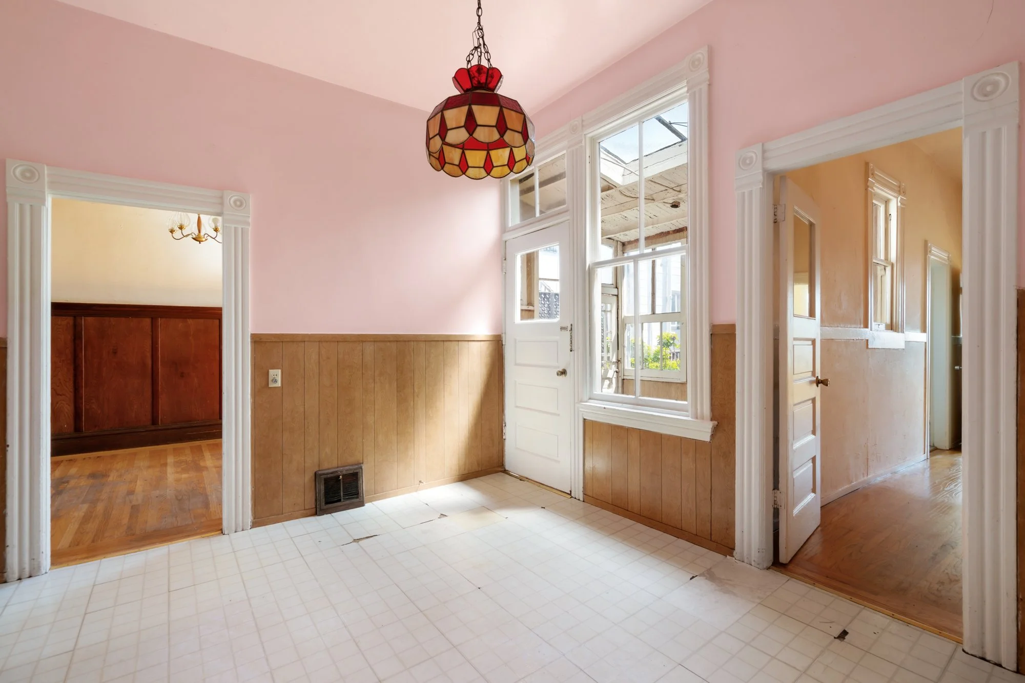 Interior view of a house showing a room with pink walls, white trim, a vintage stained glass chandelier, and large windows with white frames. Doorways lead to other rooms with darker wood paneling and walls.