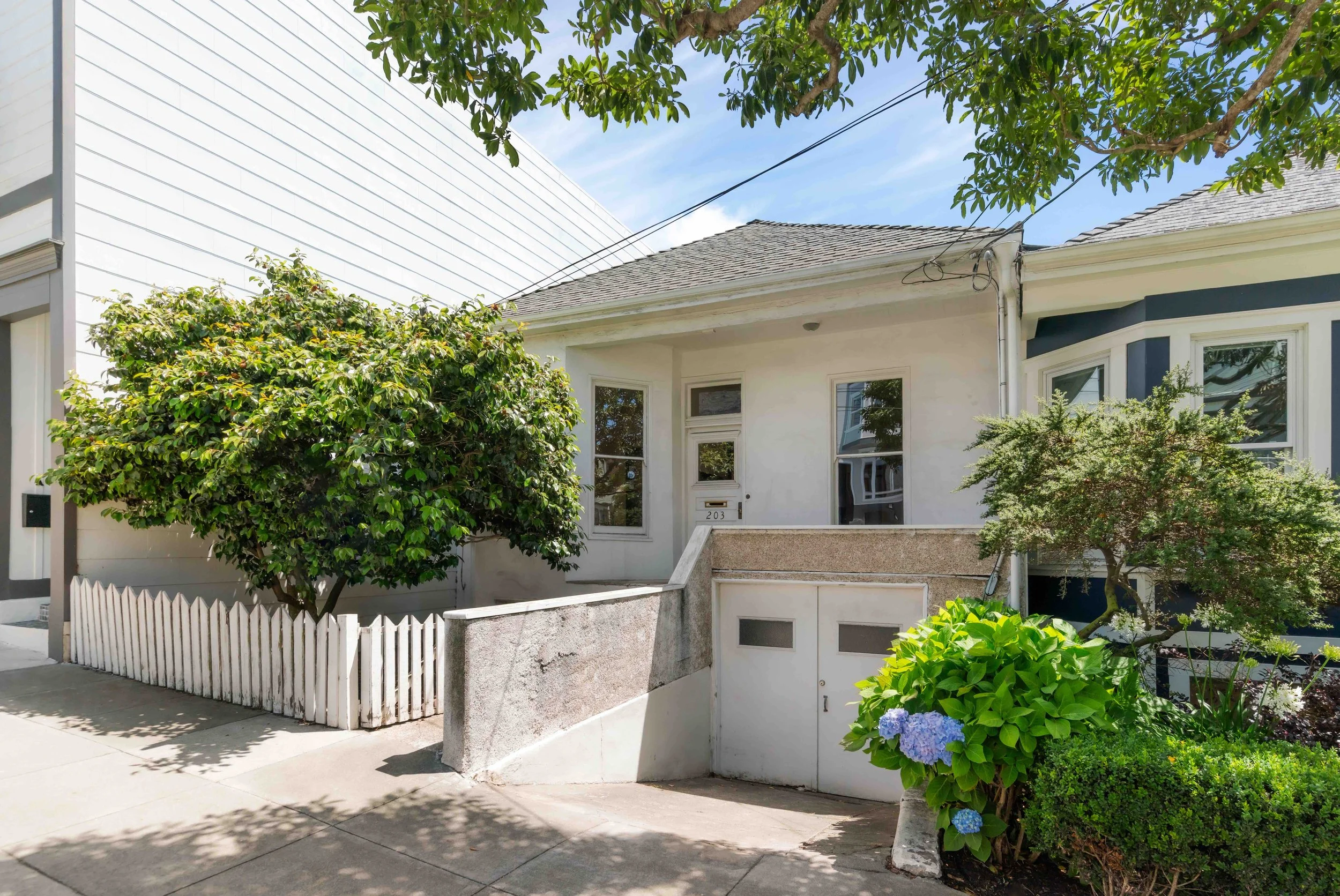 Front porch of a white house with windows, surrounded by green bushes and trees, with a small white picket fence and a sidewalk in front.