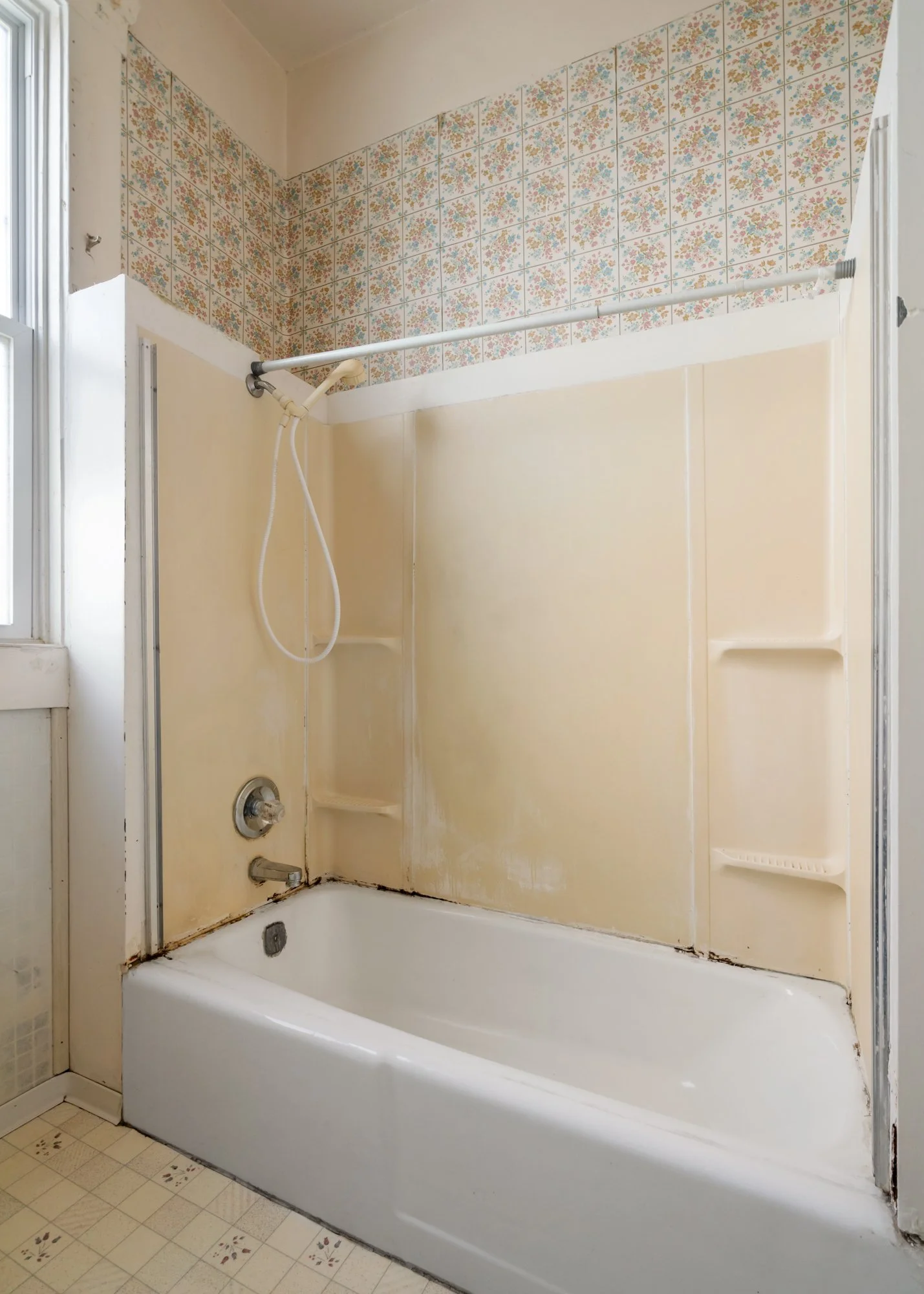 Old bathroom shower with a white bathtub, yellowish walls, floral patterned wall tiles, a window to the left, and a handheld shower head attached to the wall.