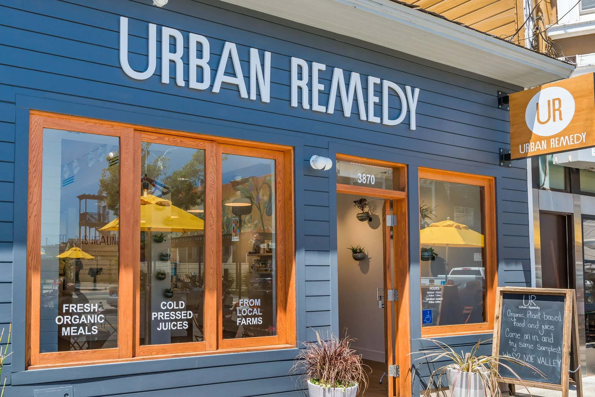 Exterior view of a storefront with blue siding and large windows, sign reading 'Urban Remedy,' promoting organic, plant-based food and juice, with yellow umbrellas and outdoor decor.