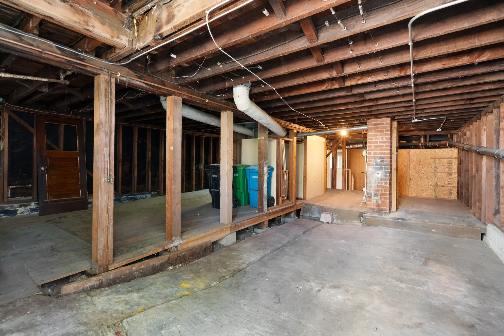 The image shows a house interior under renovation, with exposed wooden ceiling beams, electrical wires, and ducts. There are wooden studs framing parts of the walls, an industrial brick chimney, and some trash bins in the background. The floor is con