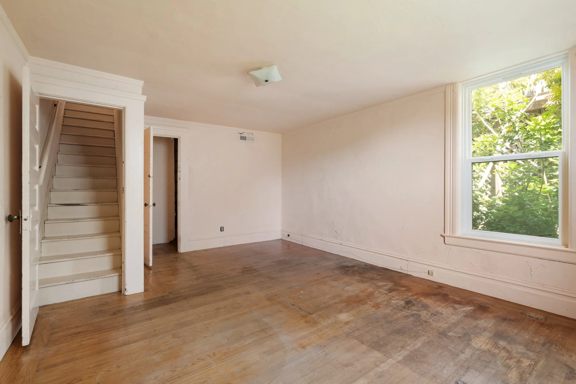 Empty living room with hardwood floors, a large window showing green trees outside, and a staircase with white risers on the left side.