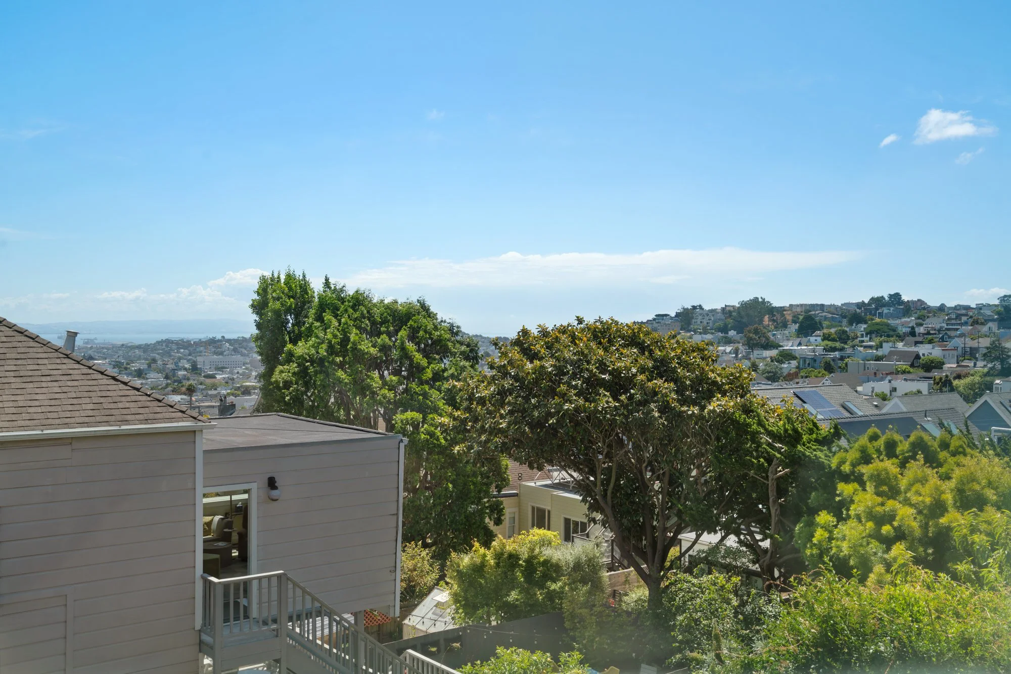 A residential neighborhood with a house in the foreground, lush green trees, and hillside houses in the background under a clear blue sky.