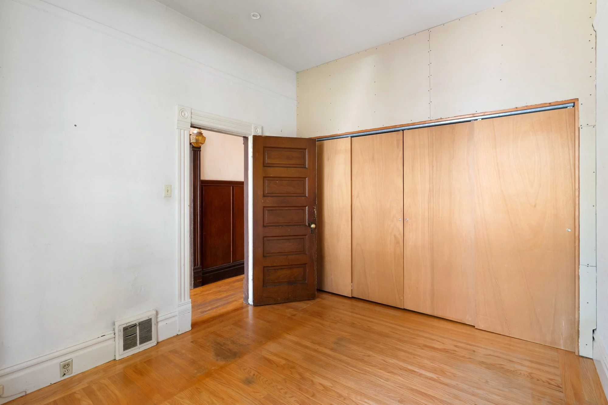 Empty room with a wooden floor, a partially open wooden door, and a large wooden closet or partition. The walls are white, and there are electrical outlets and a vent on the lower part of the wall.