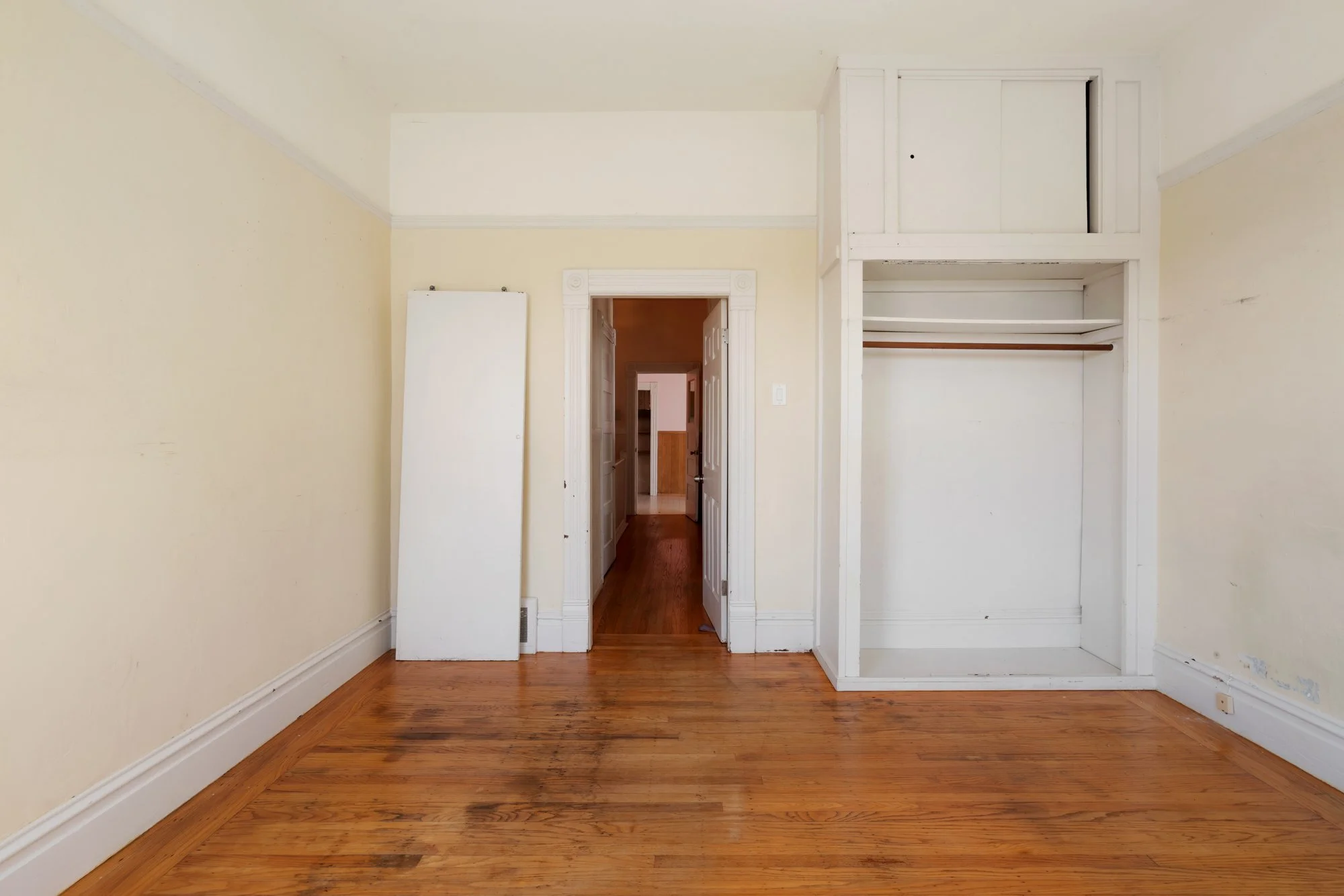 Empty room with hardwood floors, beige walls, and white trim. An open closet with a shelf and a rod, and a door leading to another room.