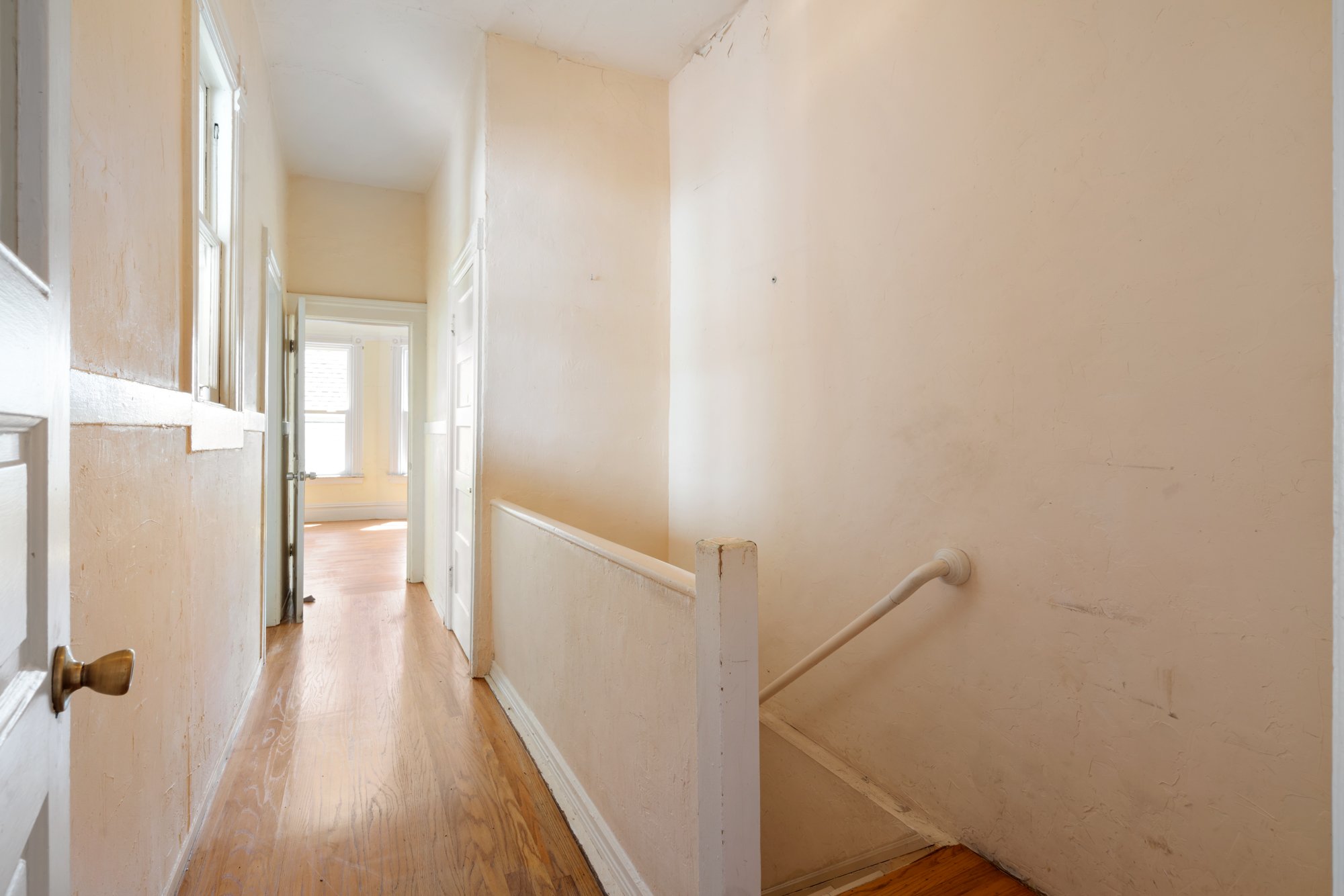 A hallway in an old house with beige walls, white trim, hardwood floors, and multiple open doors leading to rooms filled with natural light.