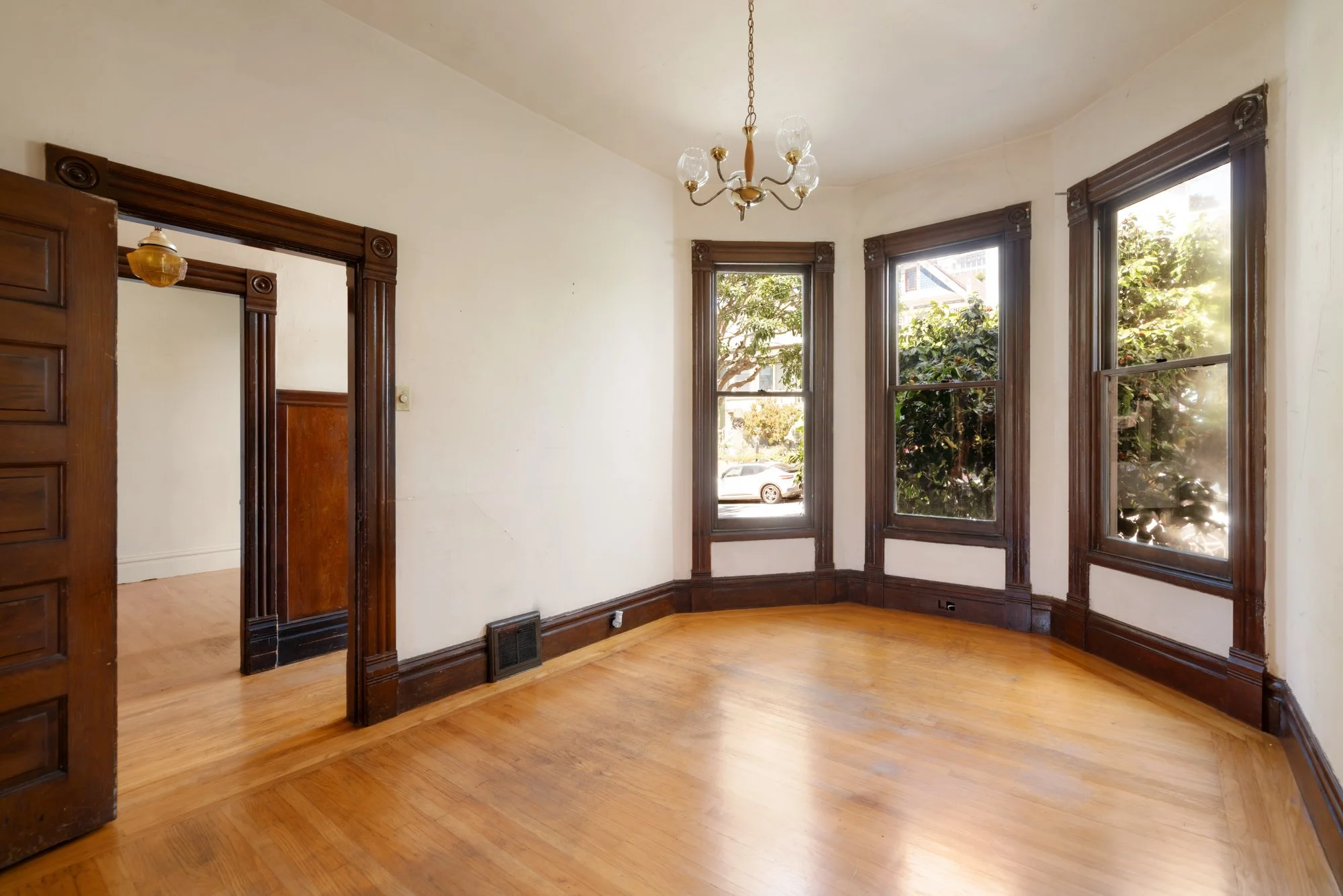 Empty room with hardwood floors, white walls, dark wooden trim around open doorways and windows, a vintage chandelier, and sunlight streaming through large windows showing trees outside.
