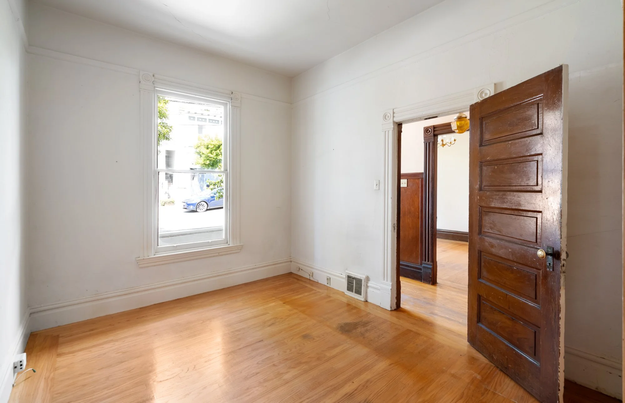 Empty room with hardwood floors, white walls, a large window, and a wooden door open to another room with wainscoting and a chandelier.