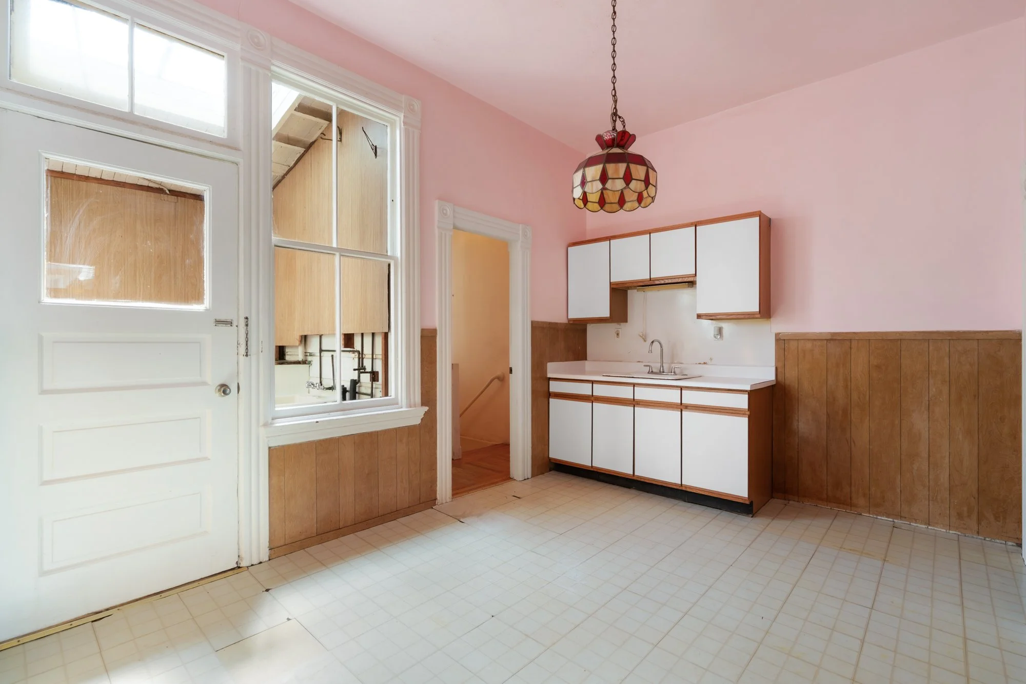 Empty kitchen with pink walls, wood paneling, white cabinets, a sink, a tiled floor, a ceiling with a stained glass light fixture, and a window with a view of an adjacent room.