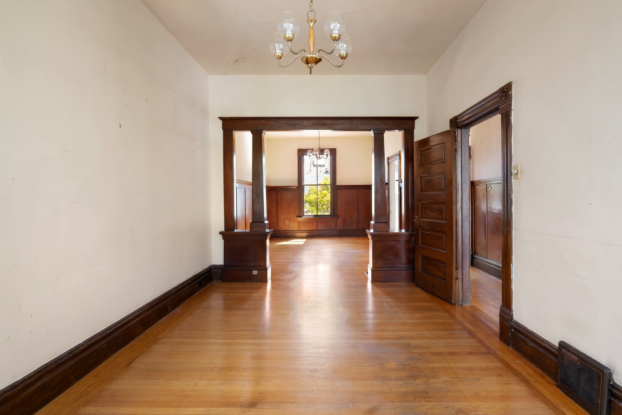 An empty room with wooden floors, cream-colored walls, and dark wood trim. There is a doorway with dark wood trim on the right, leading to another room. Through a large opening, a second room with wood paneling on the lower walls, a window, and a cha