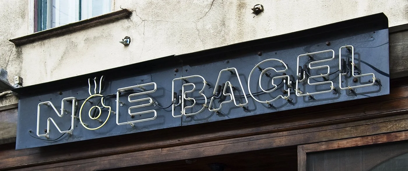 Neon sign reading 'NOEL BAGEL' on a dark background above a wooden entrance.