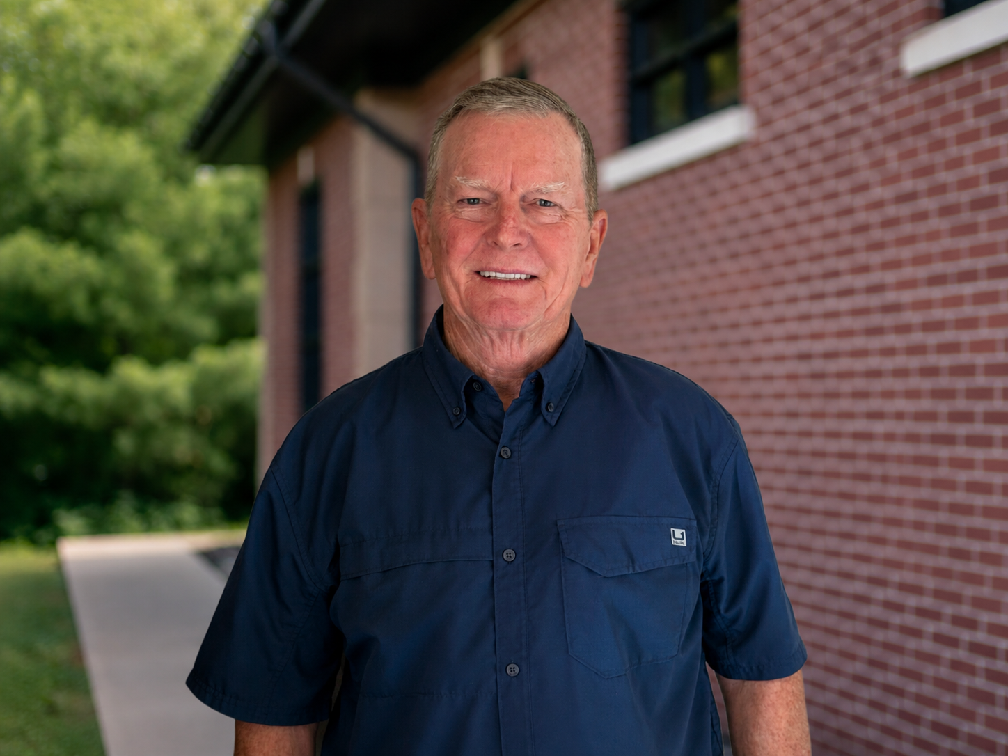 Smiling elderly man in navy blue shirt standing outdoors near a red brick building with trees in the background.