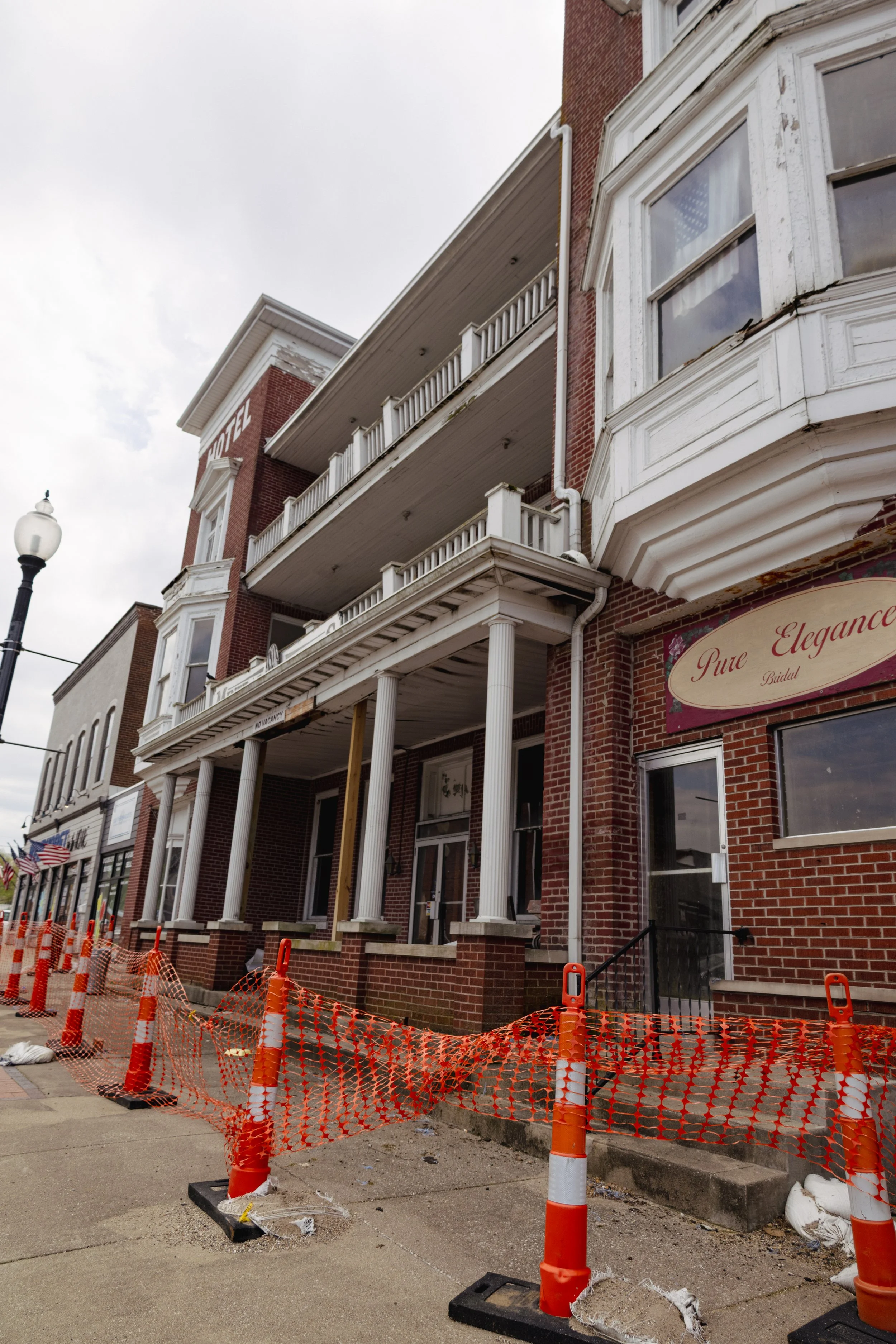 Construction site in front of Mineral Springs Hotel. Orange safety barriers and cones mark the area, with some construction materials visible. The building has white columns supporting a porch and is labeled "Hotel" on the upper part.