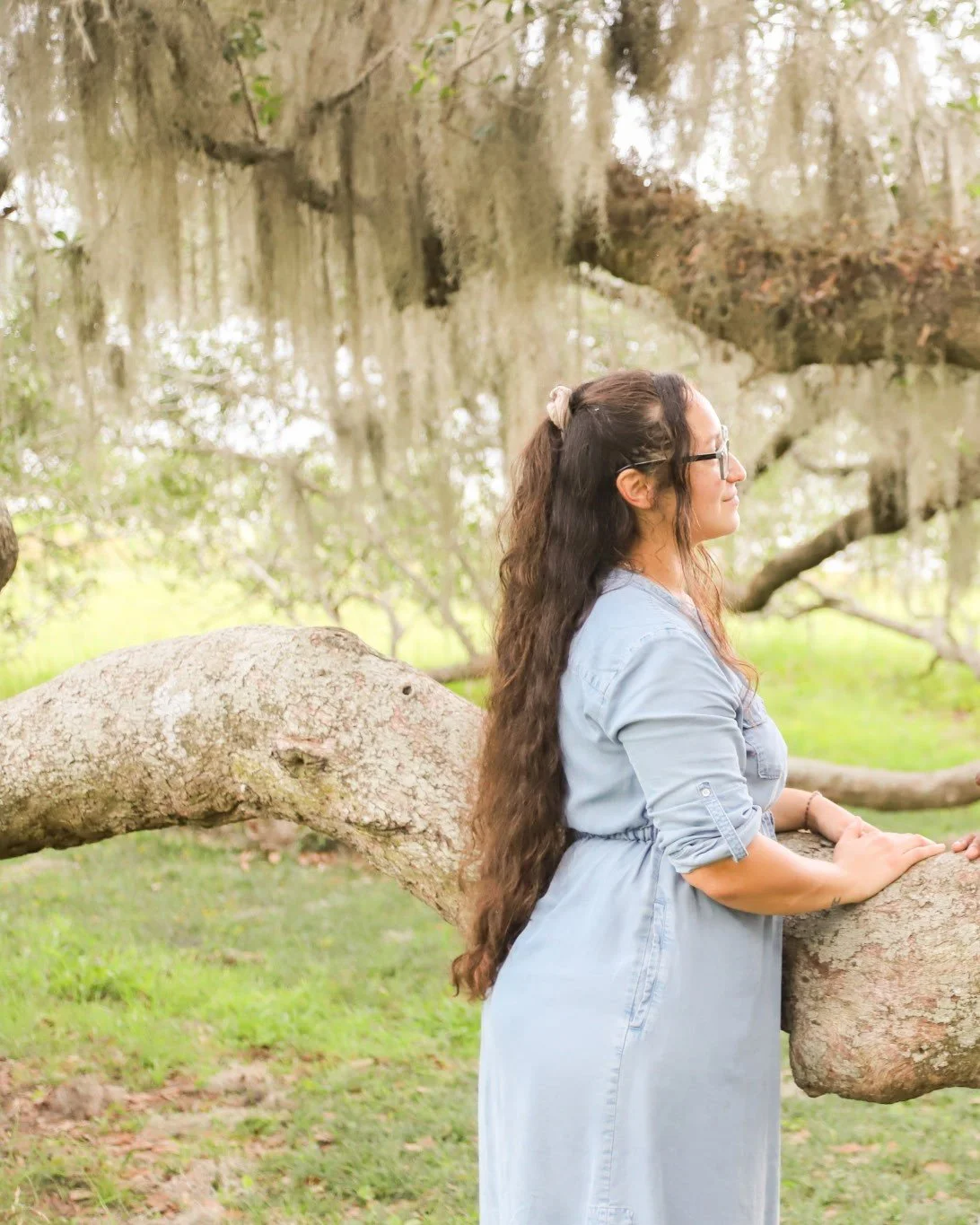 A woman with long, curly brown hair, wearing glasses and a light blue dress, standing outdoors next to a large, horizontal tree branch.