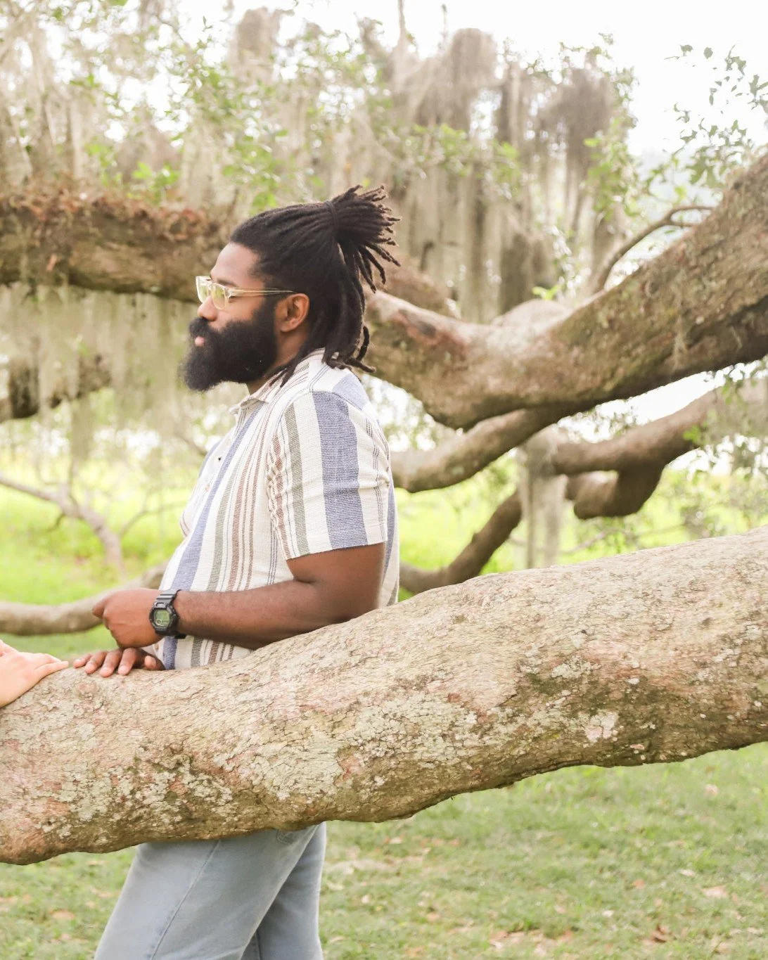A man with glasses, a beard, and dreadlocks leaning on a large tree branch outdoors in a park.