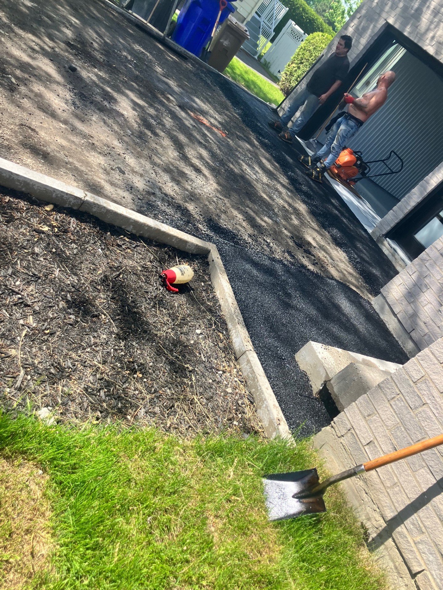 Deux hommes travaillent à la réparation ou au revêtement de la surface d'une allée ou d'un trottoir en extérieur, avec des outils et un équipement de chantier, devant un bâtiment résidentiel. Un obstacle est visible dans la zone de travail.