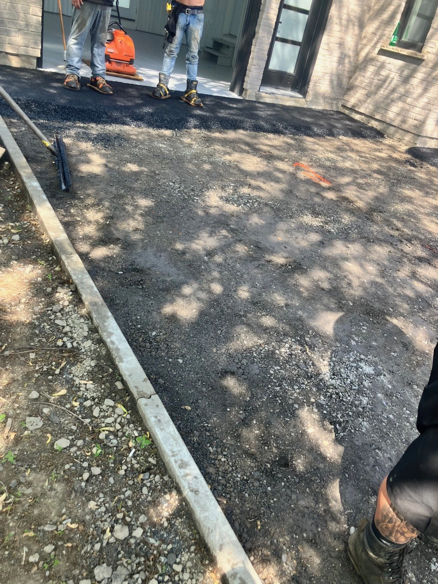 Travaux de pavage en cours devant une maison avec deux travailleurs portant des bottes de sécurité, un balai et un outil de compactage d'asphalte.