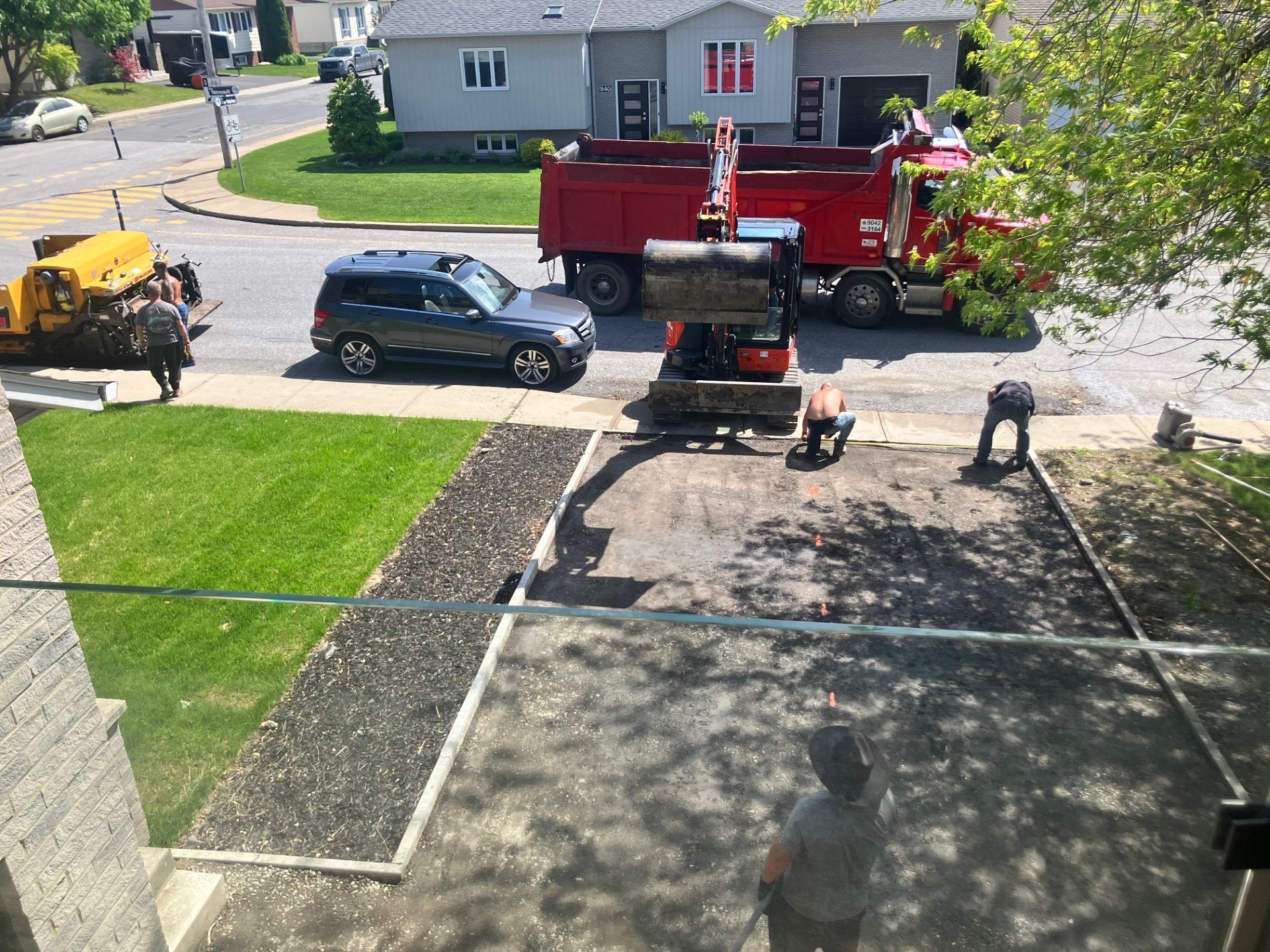 Travaux de construction ou de rénovation devant une maison, des ouvriers travaillent dans une zone délimitée, un camion rouge et une machine de construction se trouvent devant la maison, avec un véhicule noir garé à côté.