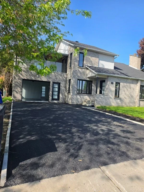 Une maison moderne avec un toit en pente, des fenêtres rectangulaires et un garage, entourée d'un espace de stationnement récemment asphalté et d'une pelouse verte, avec un arbre à gauche