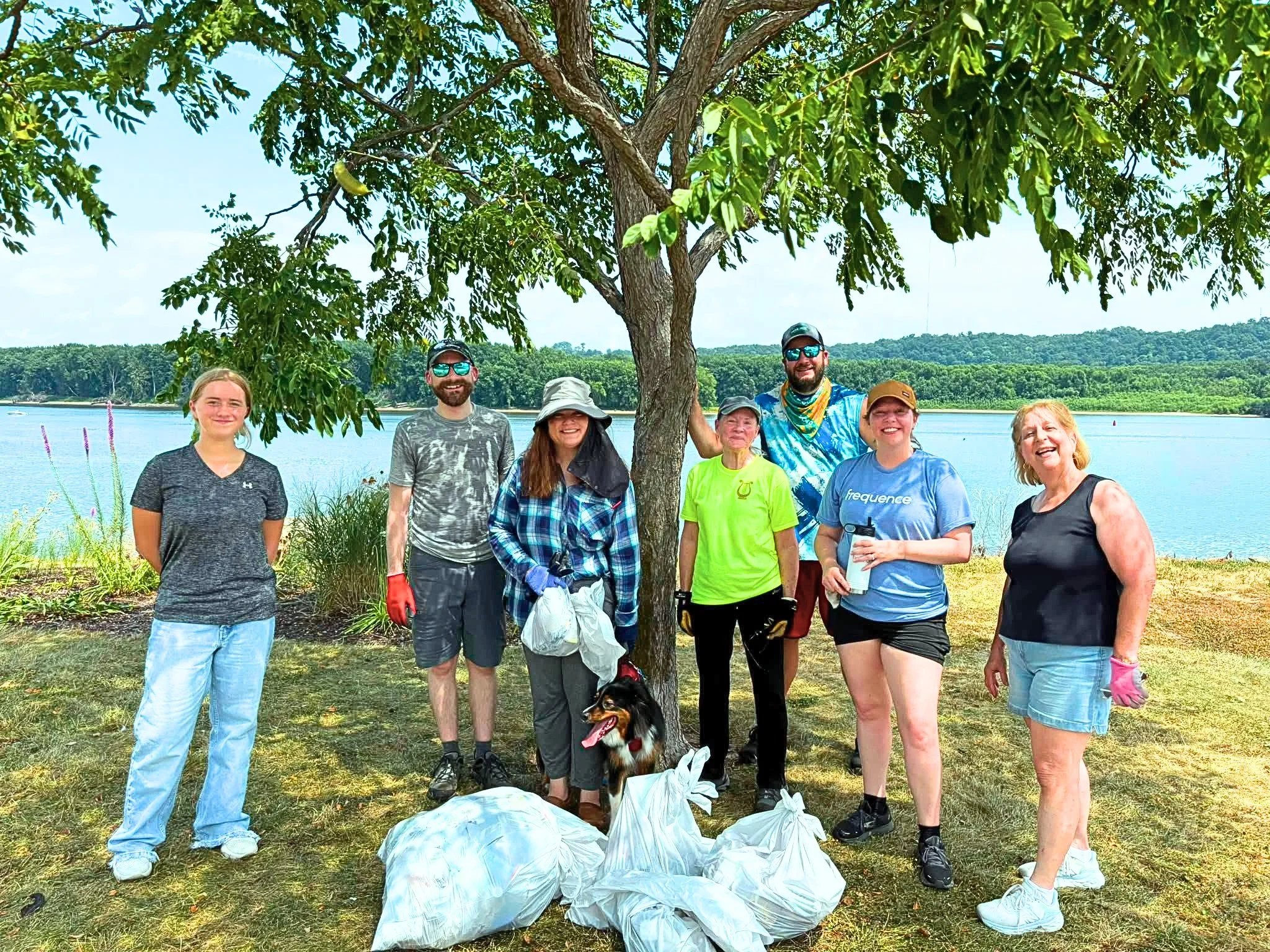 Group of seven people—including adults and children—standing under a large tree by a body of water, participating in a cleanup event with trash bags in front of them.