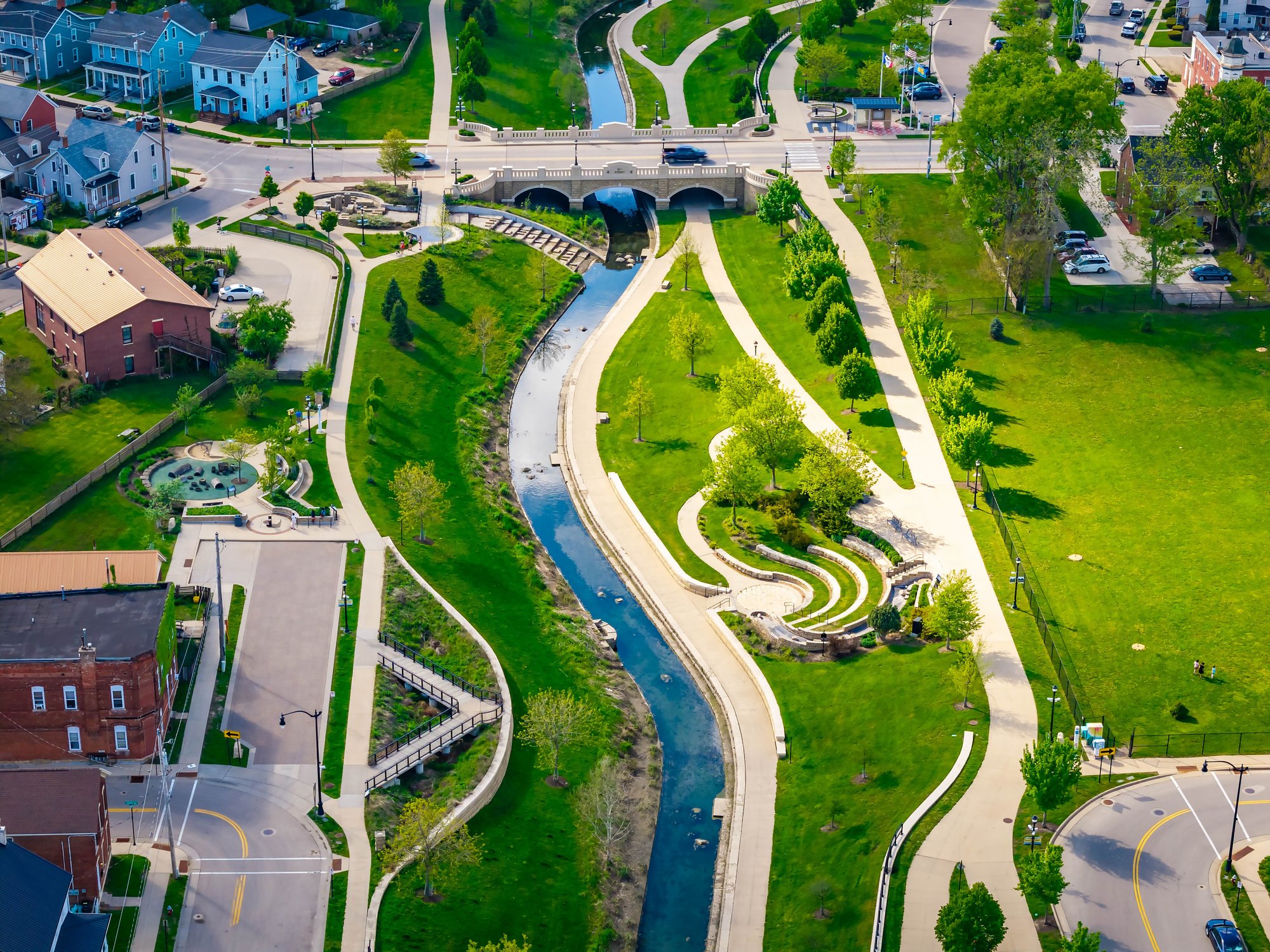 Aerial view of a park with walking paths, a small stream, and a bridge, surrounded by residential buildings and parking lots in a neighborhood.