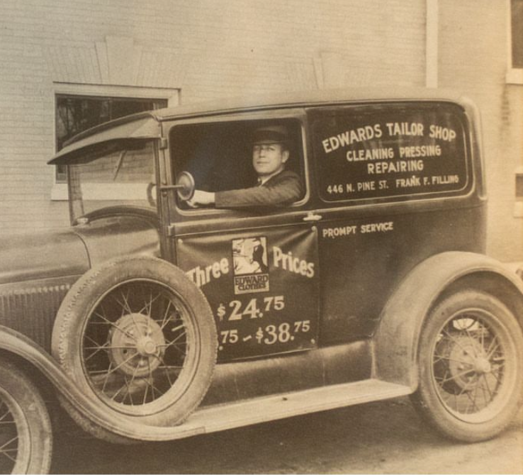 Vintage delivery truck with signage for Edwards Tailor Shop, featuring a man in the driver's seat. The sign advertises cleaning, pressing, and repairing services, and prices for three-piece suits.