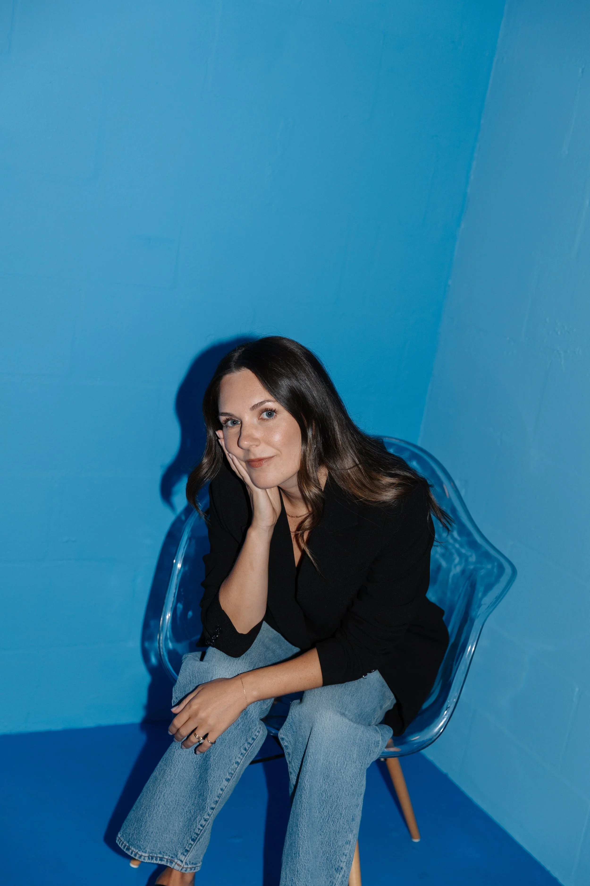 A woman with long brown hair, wearing a black blazer and light blue jeans, sitting on a clear plastic chair against a blue wall, resting her head on her hand and smiling at the camera.