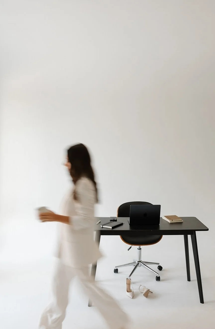 Woman walking past a black desk with a laptop, notebooks, and pens, in a minimalist white office space.