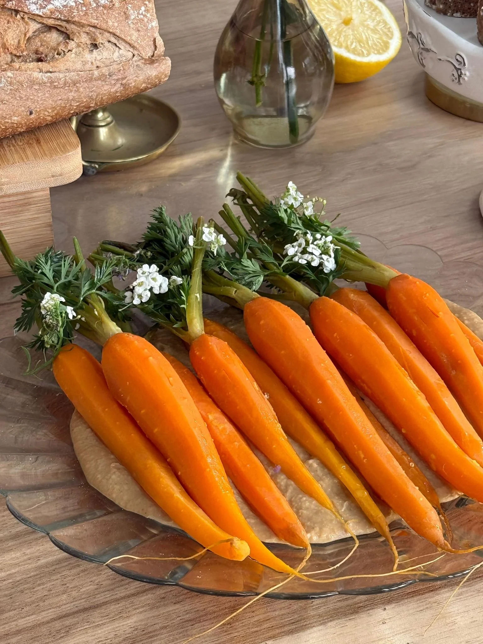 Carottes fraîches sur un plat en verre, devant une table en bois, avec un citron coupé, un vase avec une fleur et un pain dans le fond.