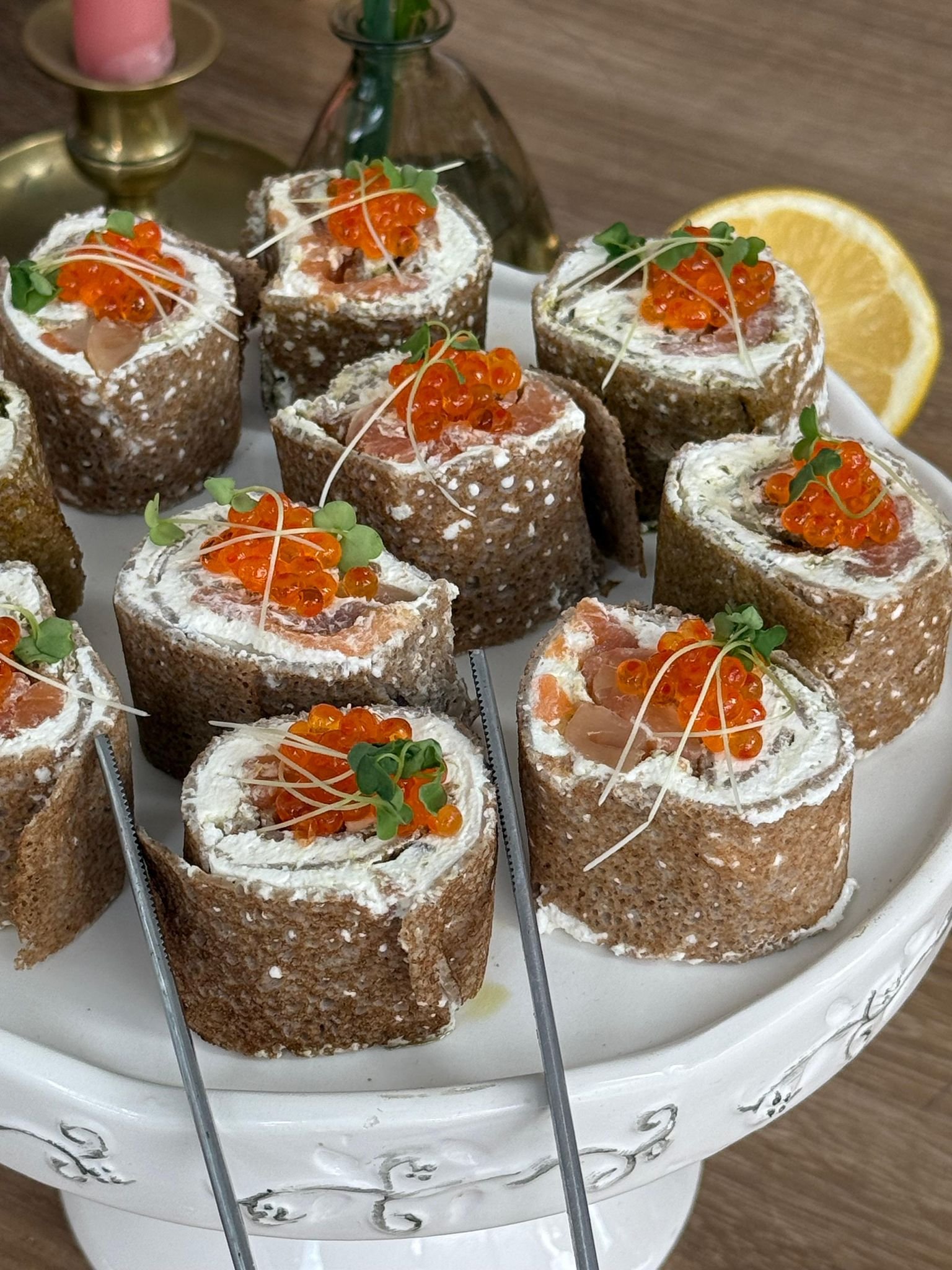 Rouleaux de saumon fumé garnis de fromage, de saumon fumé, de saumon sauvage, de câpres, de microgreens et de zestes de citron, présentés sur une assiette blanche avec un décor de fruits et plantes en arrière-plan.