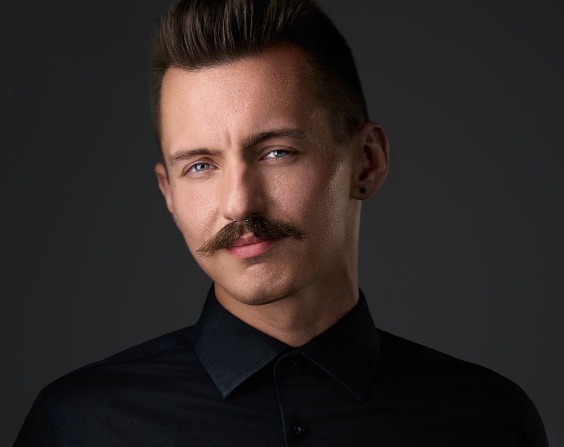 A young man with styled hair and a mustache, wearing a black collared shirt, looking confidently at the camera against a dark background.