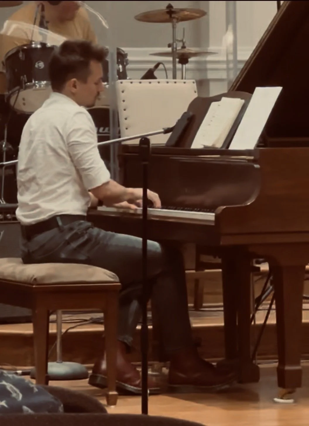 A man playing the piano in a music performance setting, with a drum set in the background.