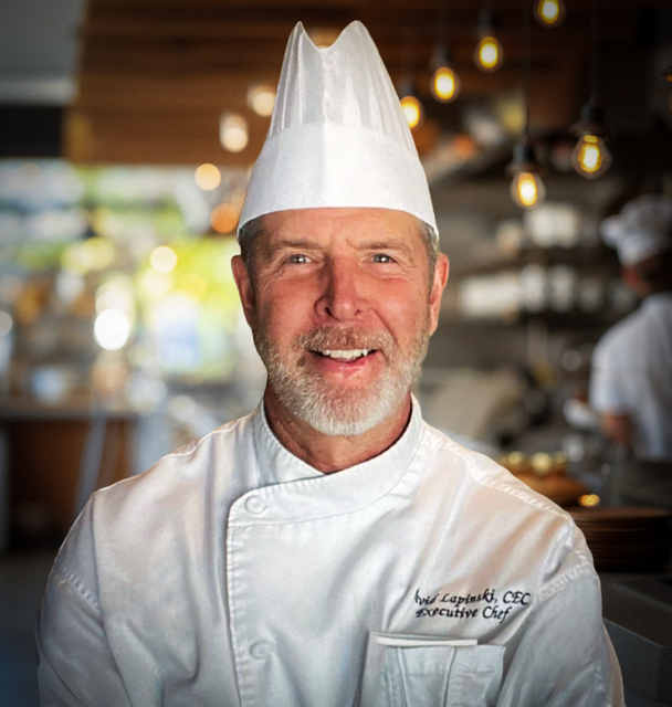 A smiling male chef in a white chef's uniform and tall white chef's hat, standing in a warmly lit restaurant kitchen.