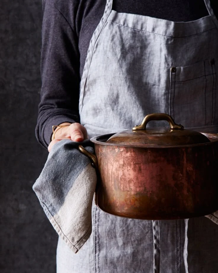 Person wearing a gray apron holding a copper pot with a cloth, standing against a dark background.